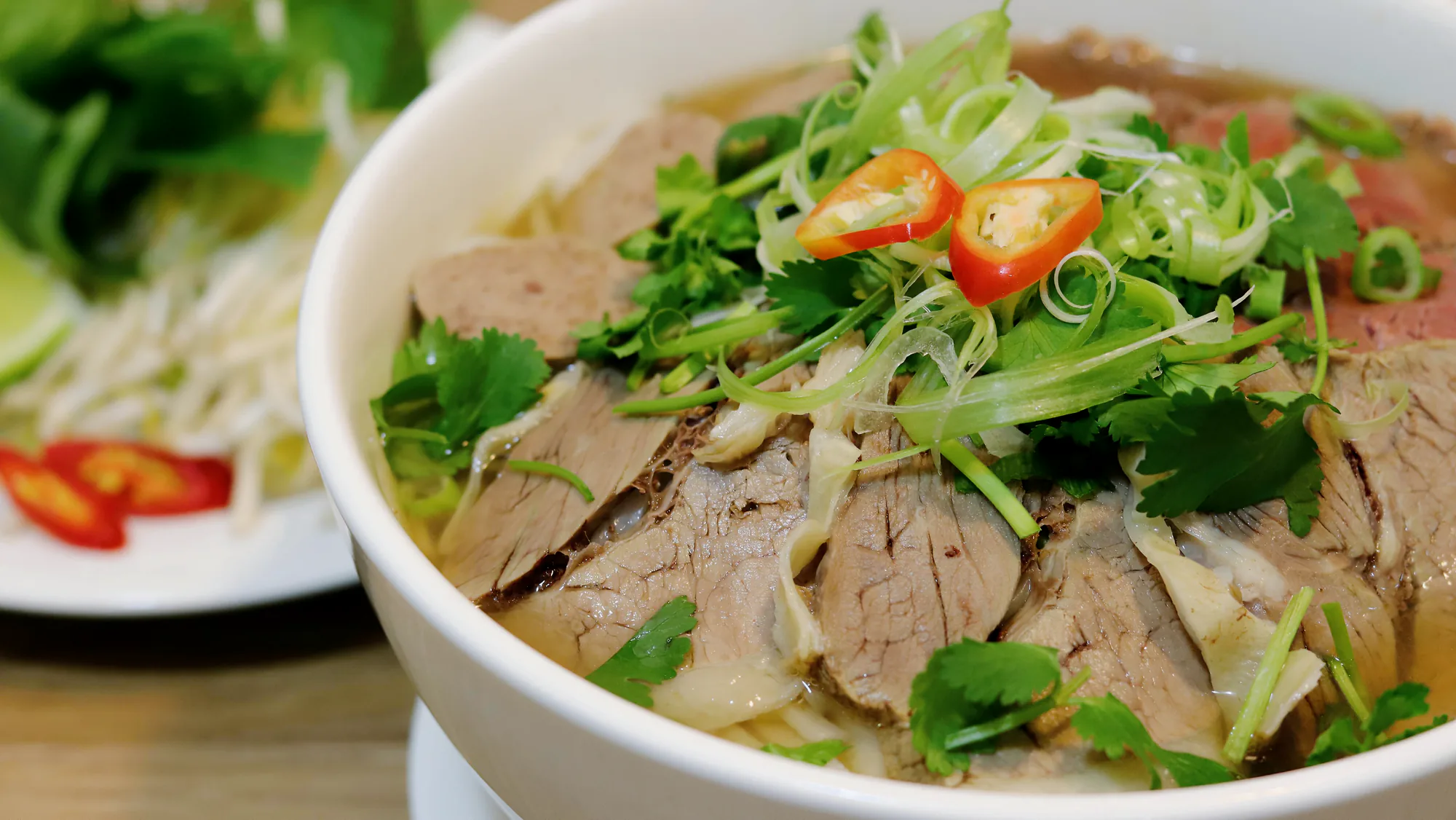 A close view of beef phở topped with herbs, spring onion and sliced chilli.