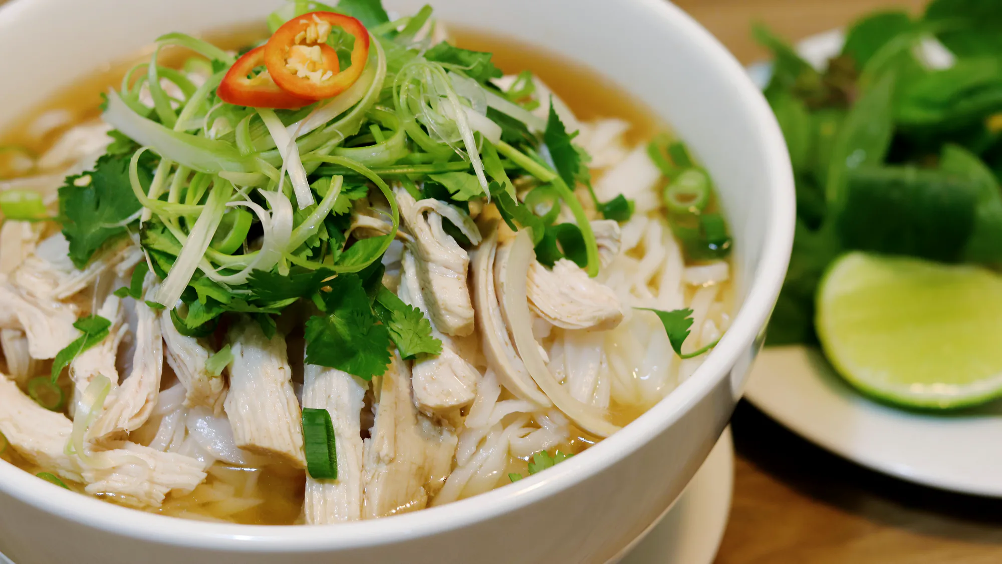 A bowl of chicken phở with herbs, spring onion, chilli and lime beside it.