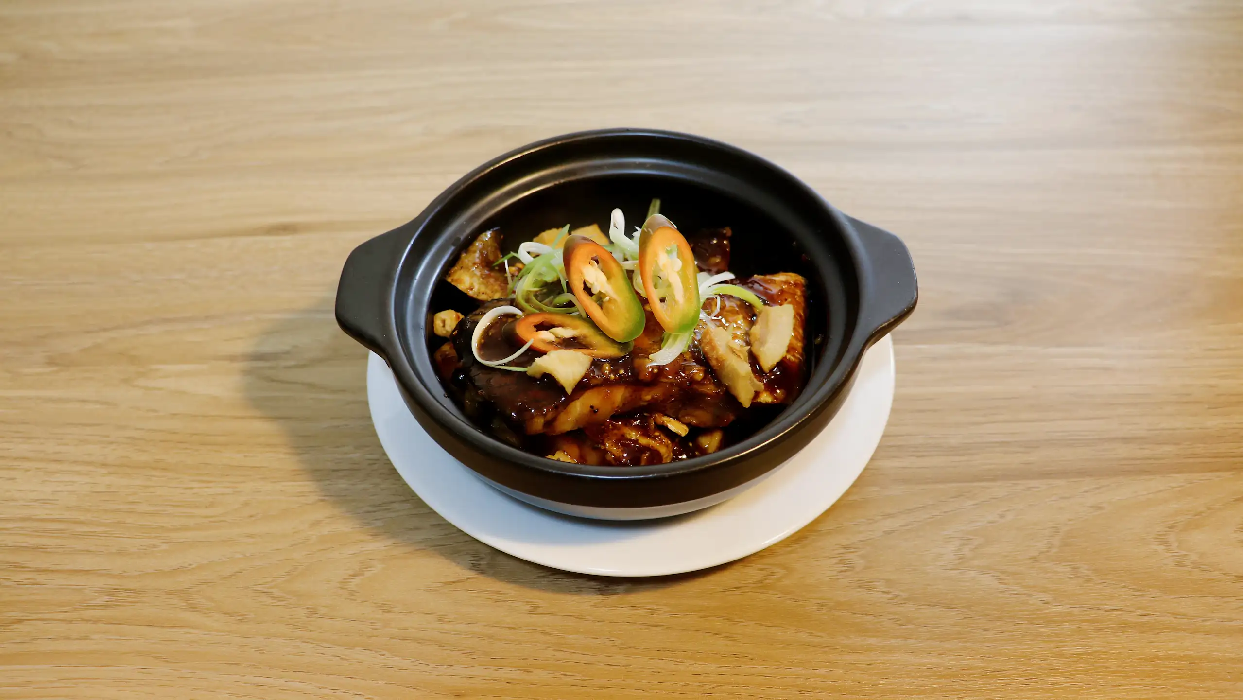 Caramelised fish pieces in a black clay pot, topped with spring onion and sliced chilli.