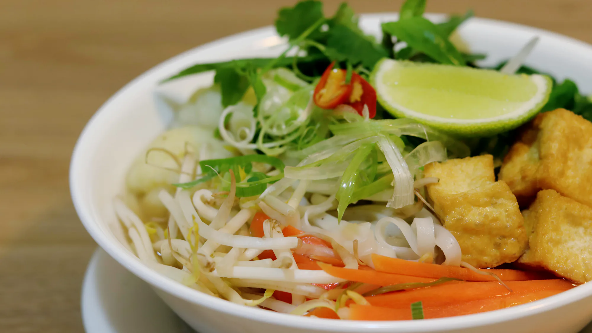 A bowl of noodle soup with tofu, bean sprouts, herbs, sliced chilli and lime.